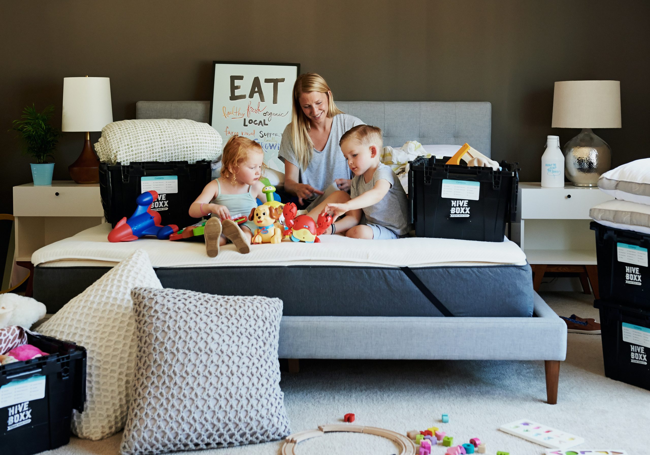 Two children playing with toys on a couch while a parent organizes moving boxes, illustrating family life during moving day.
